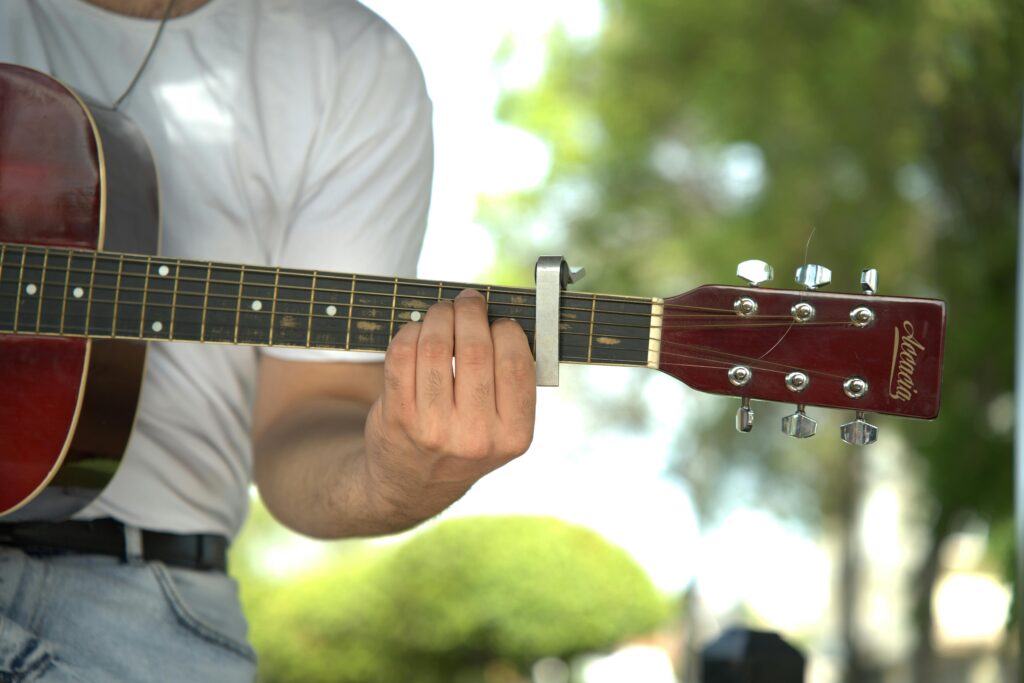 Man playing guitar outdoors with capo on fretboard, wearing casual attire.